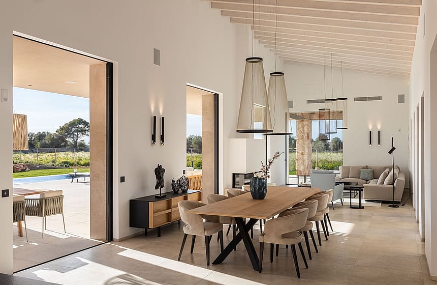 Dining room with a long oak table and cushioned chairs, open to an outdoor terrace, with white walls and exposed ceiling beams.