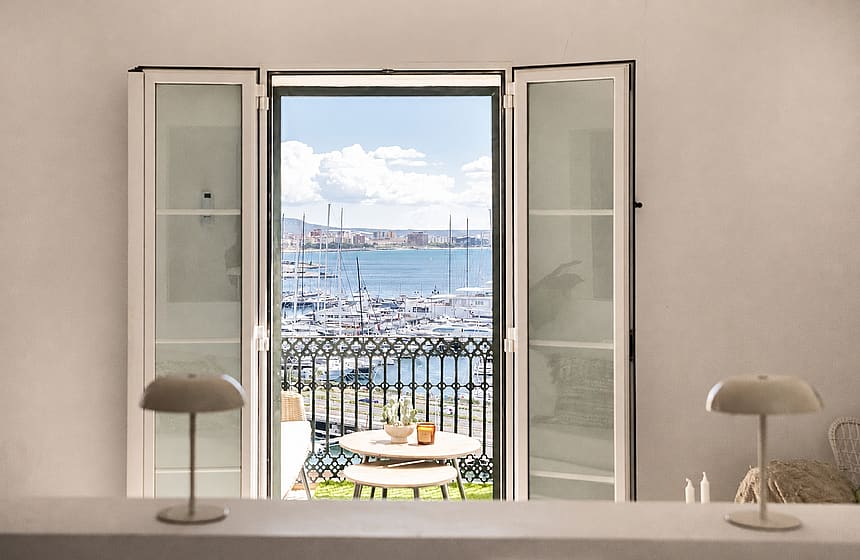 Interior view through open French doors to a small wrought-iron balcony overlooking a harbour with boats and city skyline.