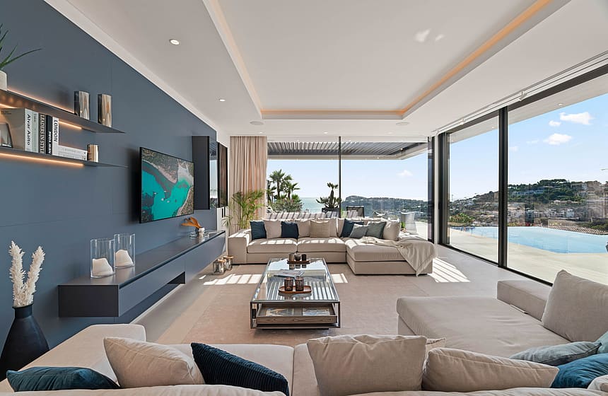 Spacious contemporary living room with a deep blue accent wall, neutral L-shaped sofas, and floor-to-ceiling glass sliding doors overlooking the sea.
