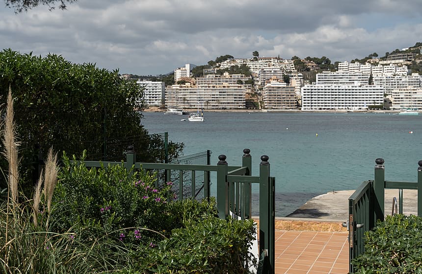 A view through a dark green garden gate over a terracotta terrace toward a calm turquoise bay and coastal buildings.