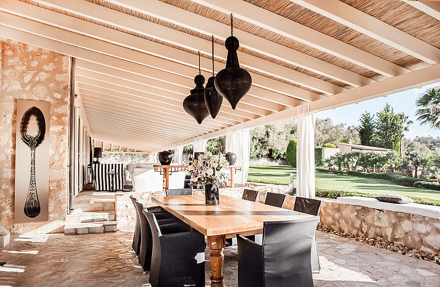A wide outdoor dining terrace featuring a long wooden table, black wicker chairs, and hanging metal pendant lights under a beamed roof.