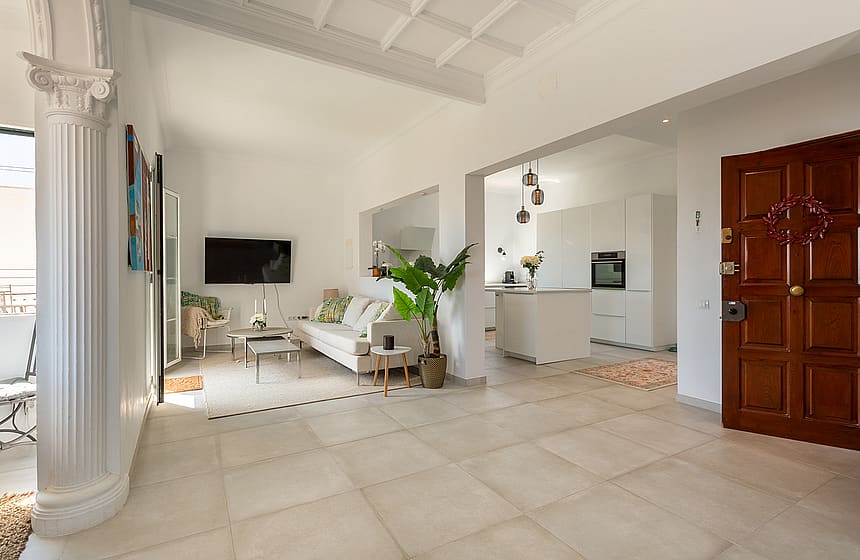 An open-plan living room in a Mediterranean apartment featuring a white column, stone tiled floors, a white sofa, and a kitchen visible in the background.