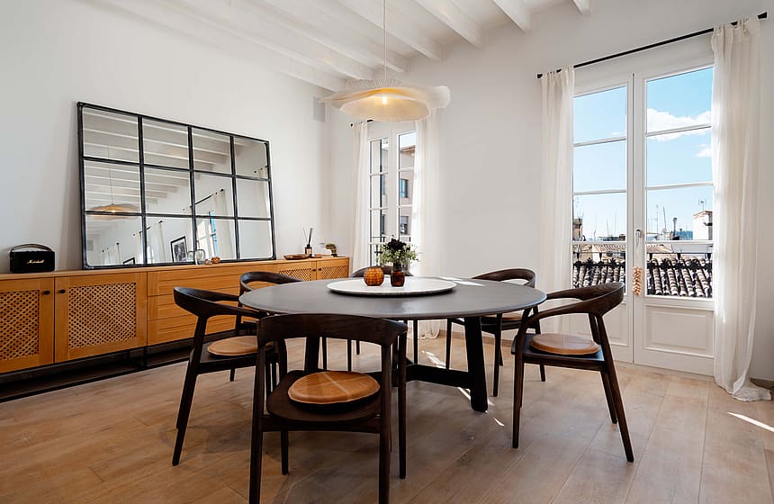 Round dark wood dining table with woven-seat chairs beneath a white beamed ceiling, French doors opening to rooftop views.