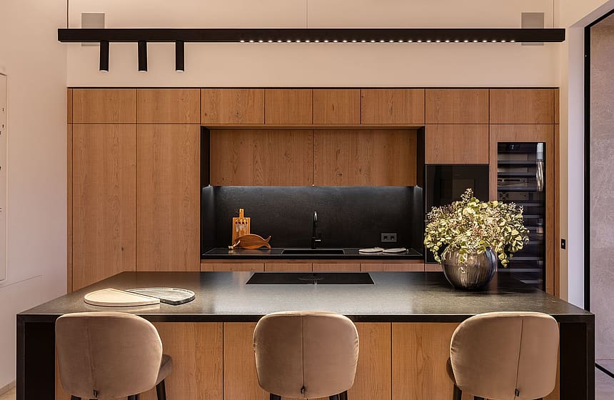 Modern kitchen featuring floor-to-ceiling oak cabinetry, a dark central island, and three beige upholstered stools.