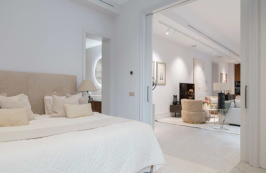 Interior view from a bedroom into an open-plan living room, featuring a beige headboard and neutral white decor.