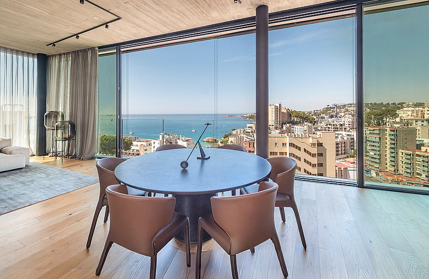 Round dark wood dining table surrounded by leather chairs next to floor-to-ceiling windows overlooking the sea and coastline.