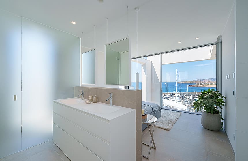 A modern master bathroom with a double vanity island, floor-to-ceiling windows, and views over Port Adriano.
