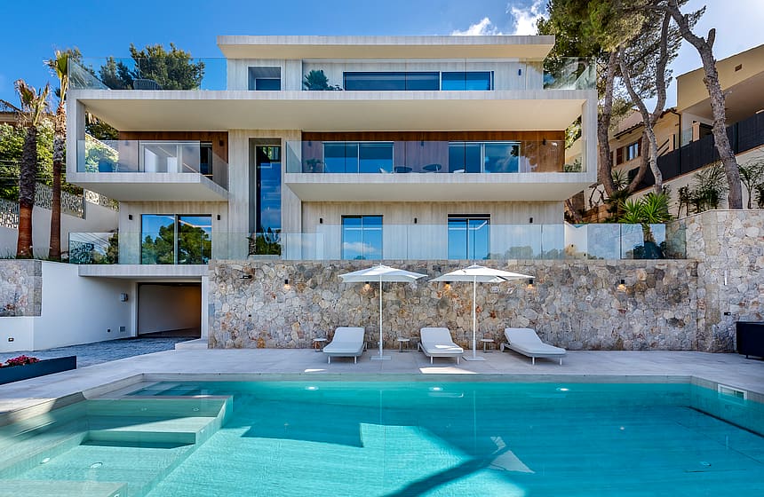 Three-story modern villa with glass railings, stone retaining wall, swimming pool, and sun loungers on a paved terrace.