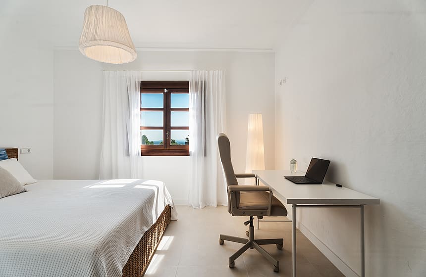 Dormitorio minimalista con cama, escritorio blanco con portátil y ventana hacia el paisaje montañoso.