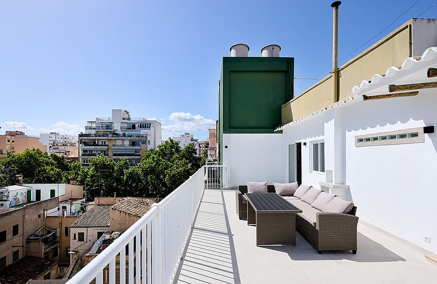 An elevated terrace with grey wicker seating and a dining table against a white exterior wall, overlooking the Santa Catalina neighborhood.