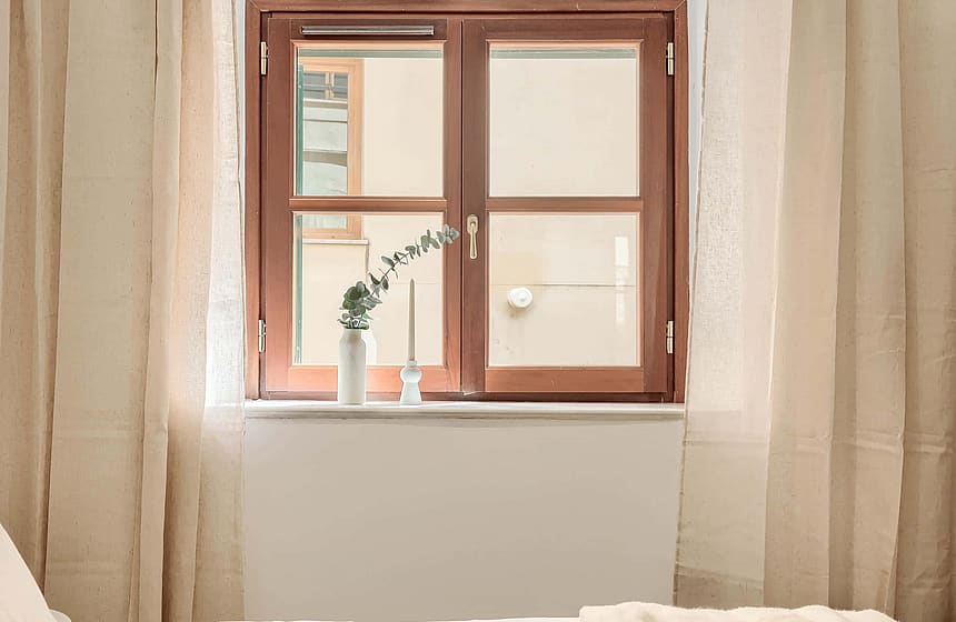 A close-up of a wooden window frame looking out onto a residential building facade, framed by floor-length linen curtains.