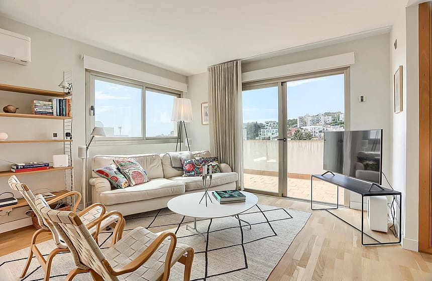 Living room featuring a beige sofa, patterned area rug, two wooden armchairs, and sliding glass doors leading to a balcony.