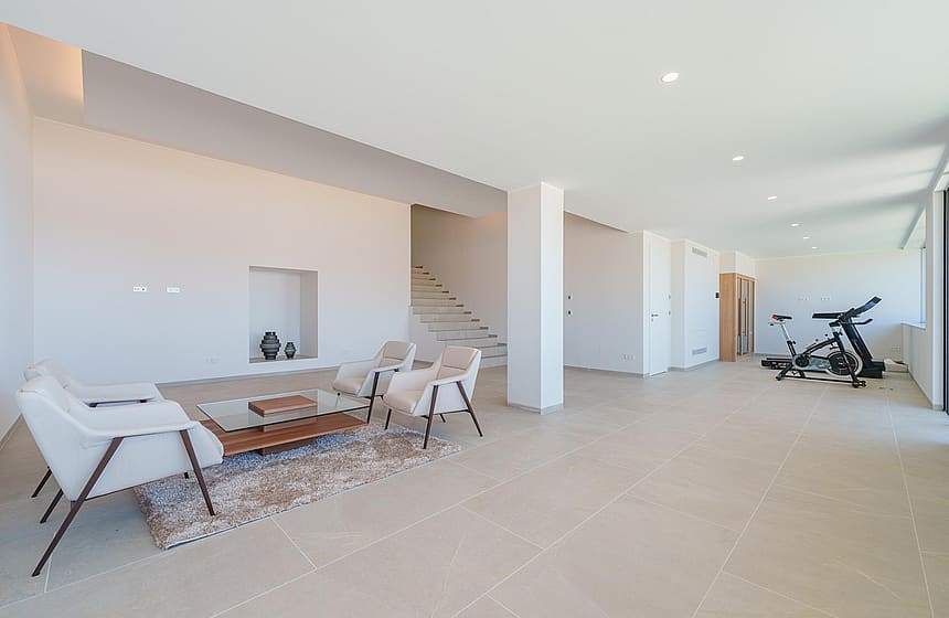 Spacious open-concept room with white walls, beige stone floors, a small lounge area, and exercise equipment in the background.