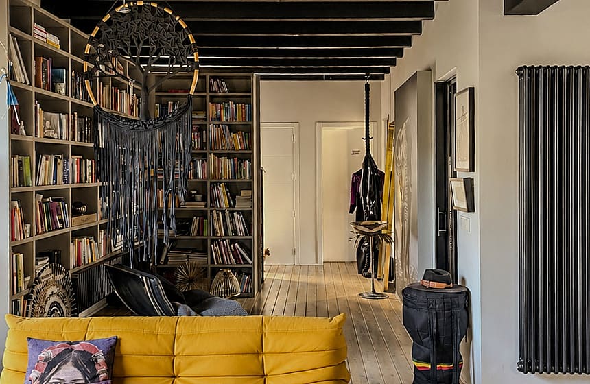 A living room interior featuring a bright yellow tufted sofa on pale wooden floorboards, dark exposed ceiling beams, and a floor-to-ceiling bookshelf.