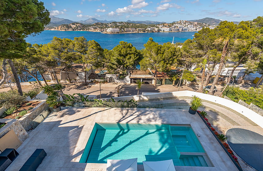 High-angle view of a rectangular swimming pool with turquoise water, set in a stone-paved terrace overlooking a bay.
