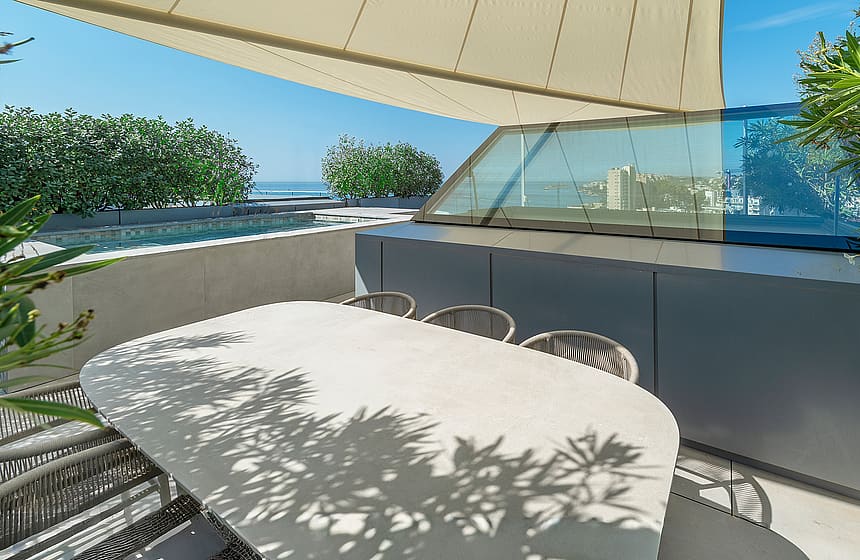 An outdoor dining table with woven chairs on a sunny rooftop terrace, featuring a glass partition and sea views.