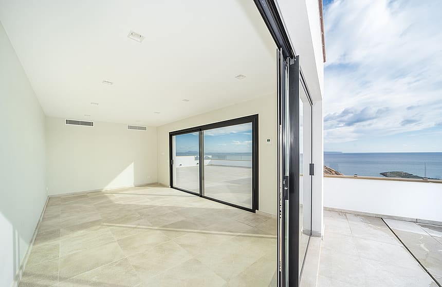 An empty modern room with light stone flooring and large sliding glass doors opening onto an outdoor sea-view terrace.