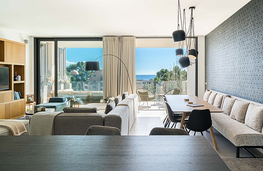 Open-plan living and dining room with a long beige bench seat, dark pendant lights, and floor-to-ceiling balcony views.