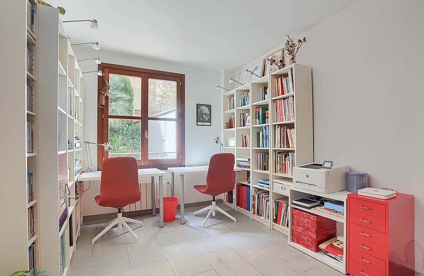 A home office featuring two white desks, red rolling chairs, and floor-to-ceiling white shelving units under a wooden window.