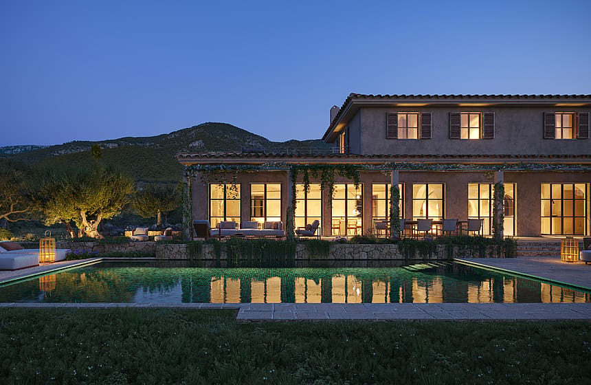 Exterior view of a stone finca at dusk, featuring a rectangular swimming pool, illuminated ground floor windows, and surrounding mountain silhouettes.
