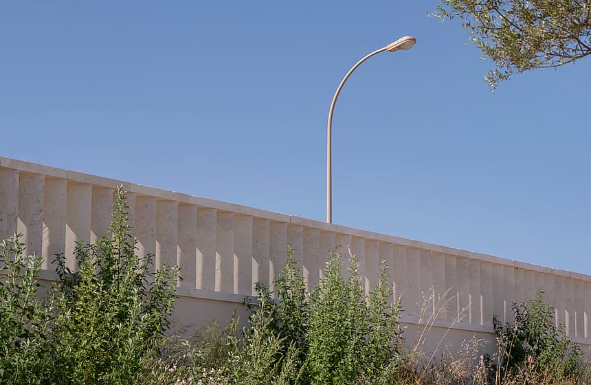A beige stone wall featuring vertical recessed slats stands under a clear blue sky with a curved metal street lamp.