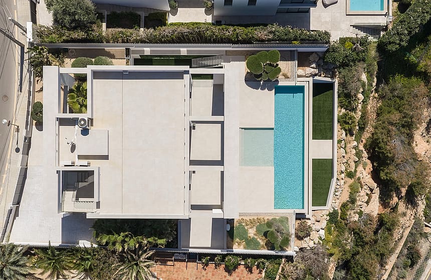 Top-down drone shot of a modern white villa with a rectangular swimming pool, terraced gardens, and surrounding Mediterranean landscape.
