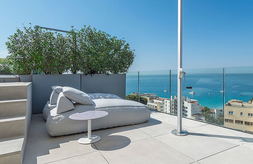 A rooftop terrace featuring a large gray outdoor daybed, a white side table, and a glass balustrade overlooking the sea.