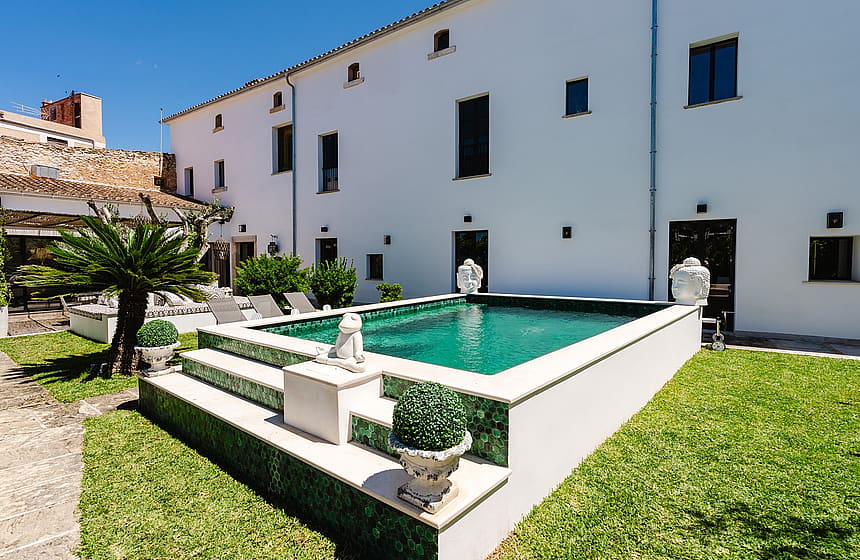 A raised outdoor swimming pool with green tiled edges and stone steps, set in a lawn next to a white traditional townhouse facade.