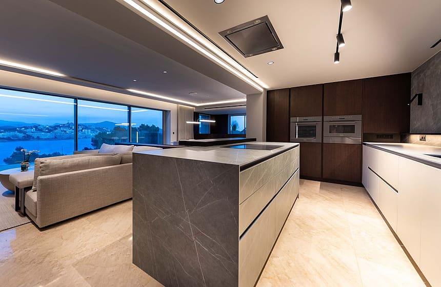 A contemporary kitchen featuring a stone-topped central island, dark wood cabinetry, and integrated appliances under recessed lighting.