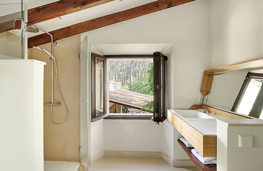 A bathroom in a finca with white walls, wooden ceiling beams, a floating vanity, and an open window showing trees.