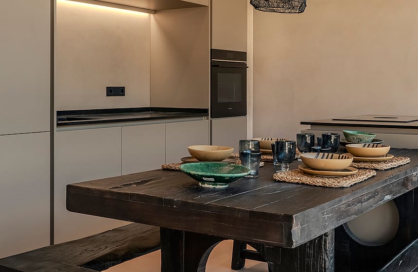A modern kitchen featuring floor-to-ceiling beige cabinets and a dark, weathered timber dining table with matching bench seating.