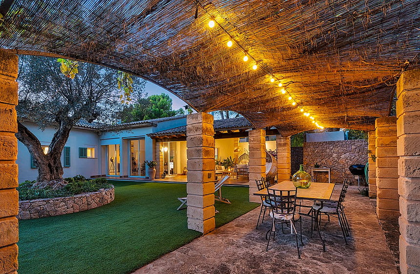 A covered terrace with natural stone pillars and a reed roof, featuring an outdoor dining area with wrought iron chairs, overlooking a landscaped garden with an olive tree at dusk.