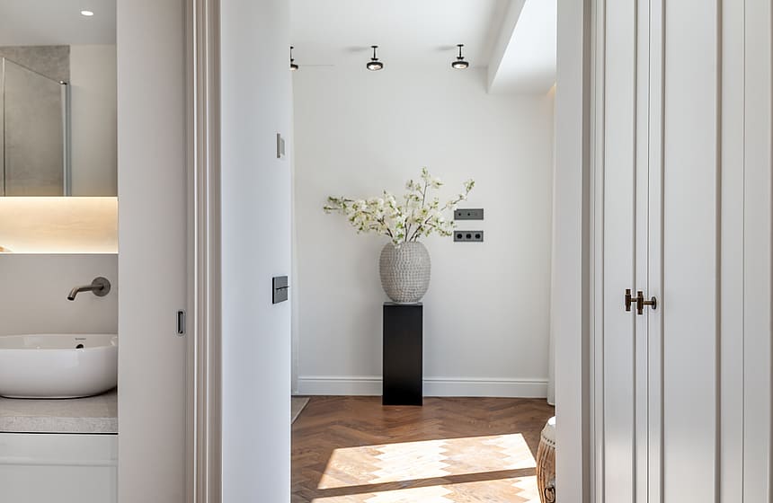 A view through a doorway showing herringbone oak floors, white walls, and a black pedestal with a floral arrangement.