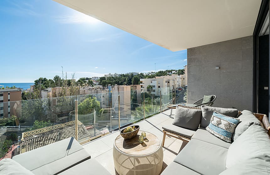 A covered terrace with grey outdoor sofas, a circular wooden coffee table, and glass railings overlooking a coastal town.