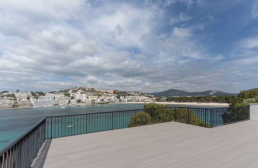 A wide grey-decked terrace with black metal railings overlooking a Mediterranean coastal town, sandy beach, and mountains.