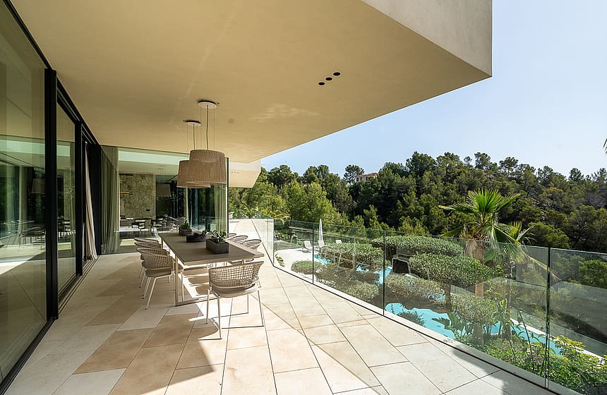 An expansive covered outdoor terrace featuring a dining table, natural stone flooring, and a glass balustrade overlooking trees.