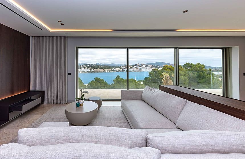 Modern living room with a long beige fabric sectional sofa facing floor-to-ceiling glass windows overlooking a blue bay.