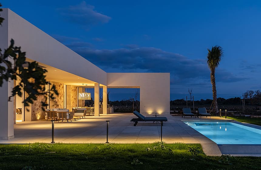 Exterior view of a modern finca at twilight, featuring a lit swimming pool, stone terrace, and warm architectural lighting.