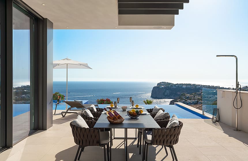 Covered outdoor dining terrace with a dark metal table, woven chairs, and an infinity pool edge against a Mediterranean sea view.