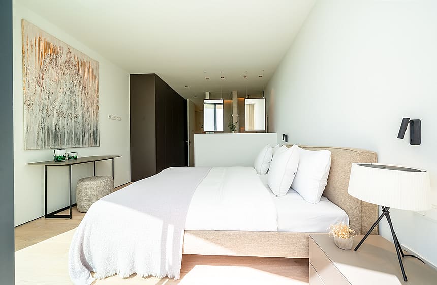 A modern master bedroom with a king-sized bed, white linens, a textured headboard, a console table with abstract art, and an open en-suite bathroom area.