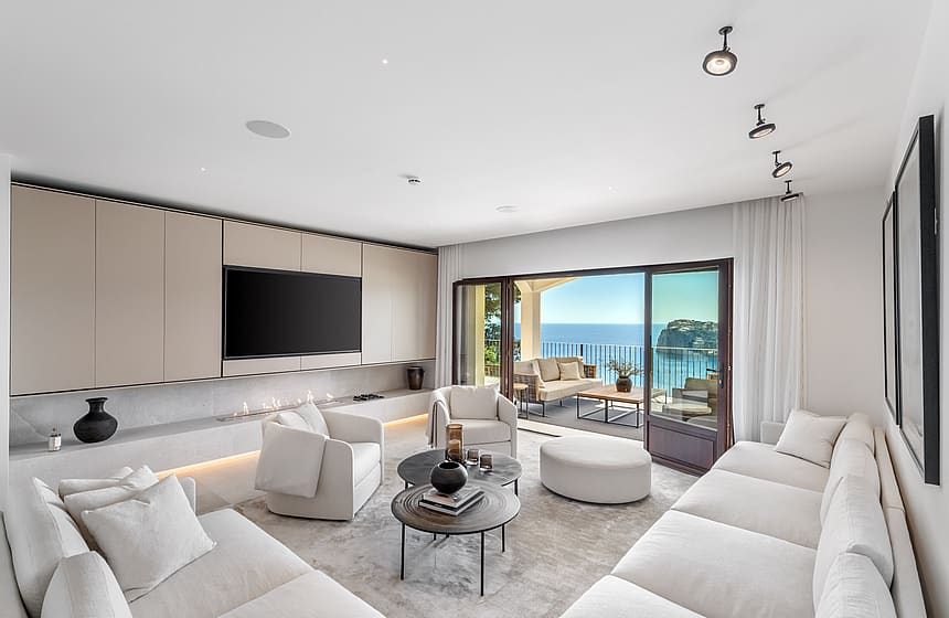 Modern white living room with soft seating and circular tables facing wide glass doors and a coastal view.