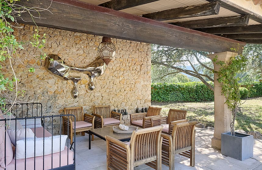 A shaded outdoor terrace with a natural stone wall, teak furniture, and a metal sculpture, overlooking greenery.