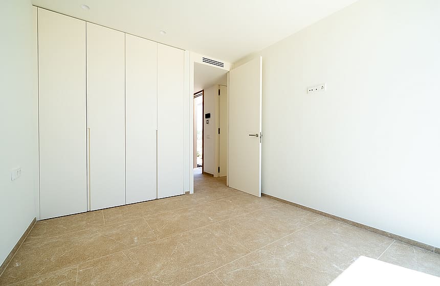 An empty bedroom featuring white built-in closets, plain walls, and beige carpet flooring with a door opening to a hallway.
