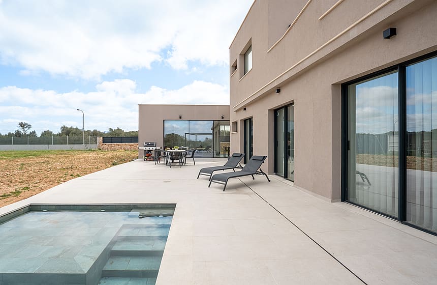 Wide view of a modern villa terrace featuring a rectangular swimming pool, sun loungers, and large sliding glass doors.