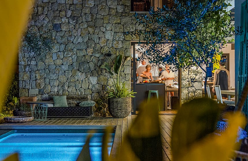 A stone-walled outdoor terrace with a swimming pool and a view into a lit interior kitchen, framed by foreground foliage.