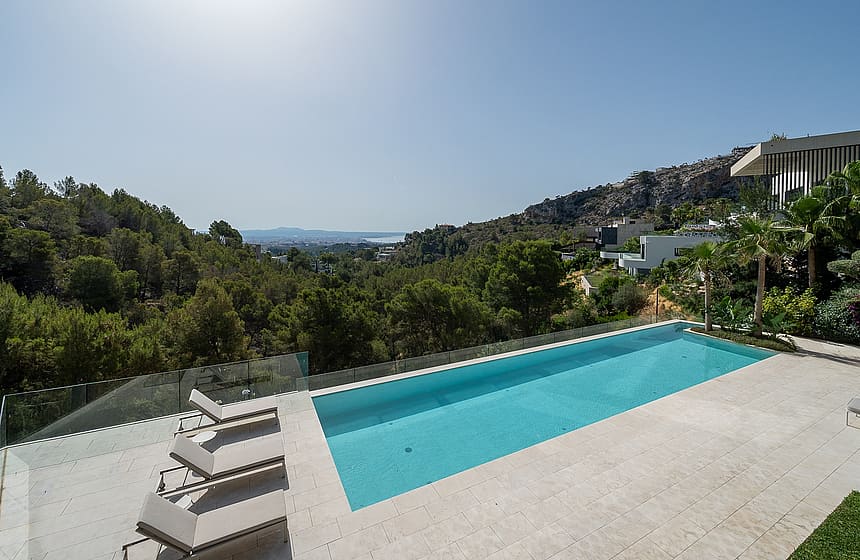 Rectangular swimming pool with clear blue water and stone deck, set against a backdrop of forested hills and distant sea.