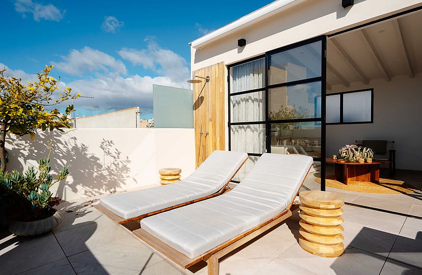 A private terrace in Santa Catalina featuring two wooden lounge chairs, a wall-mounted shower, and a black-framed glass door.