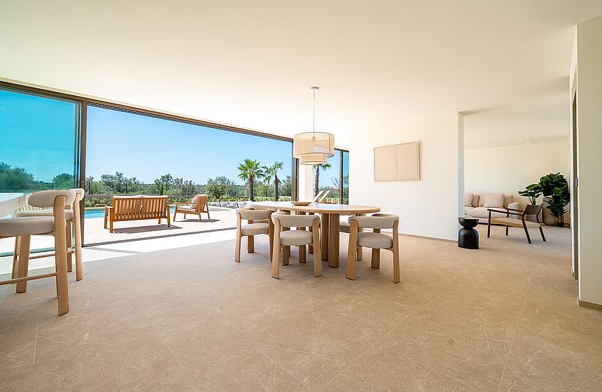 Dining room with circular wooden table and upholstered chairs, opening to a terrace with mountain views in Son Gual.