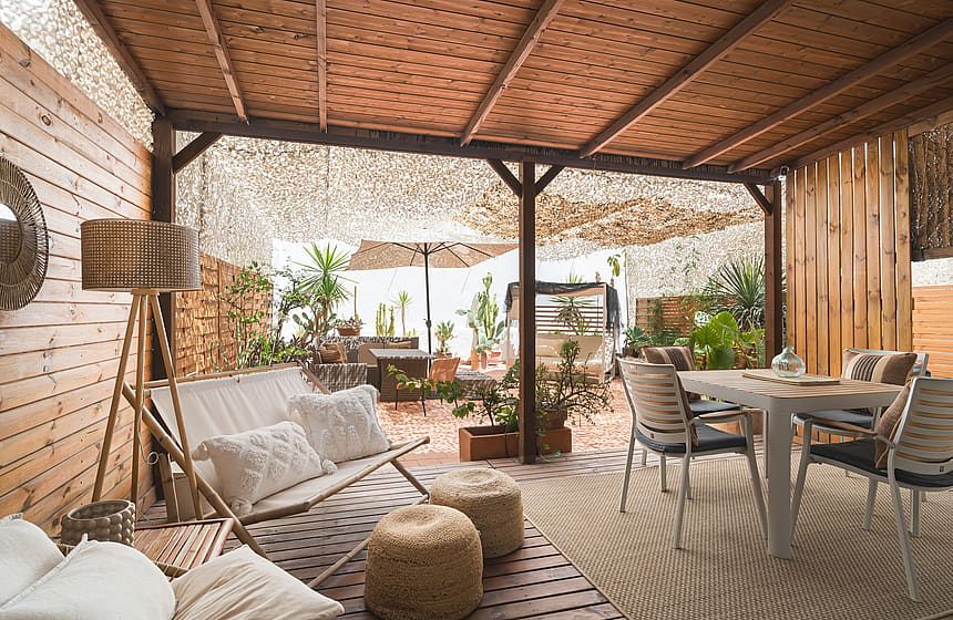 A shaded outdoor terrace with wooden panel walls, dining furniture, a sofa, and potted plants under a perforated roof.