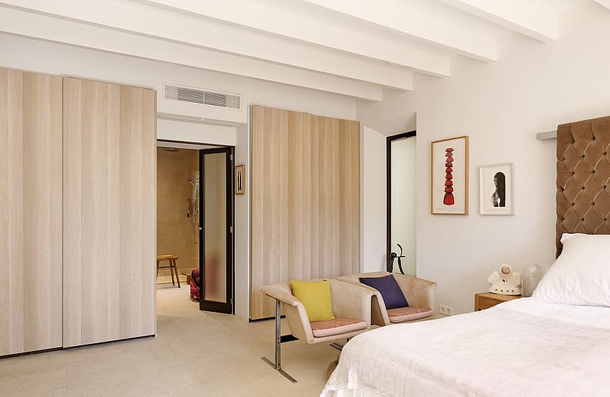 A bright bedroom with white beamed ceiling, light wood wardrobe doors, and a partial view into an en-suite bathroom.